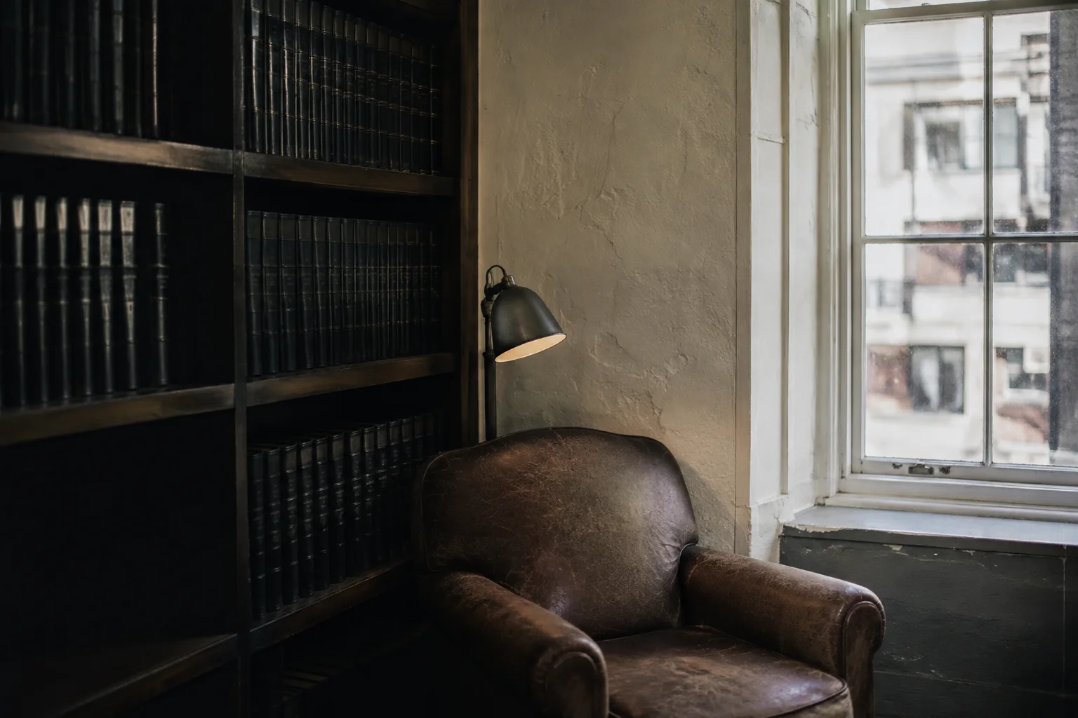 An independent law office reference room with shelves of unlettered dark bound volumes, a worn leather chair and small reading lamp, pale plaster and daylight.