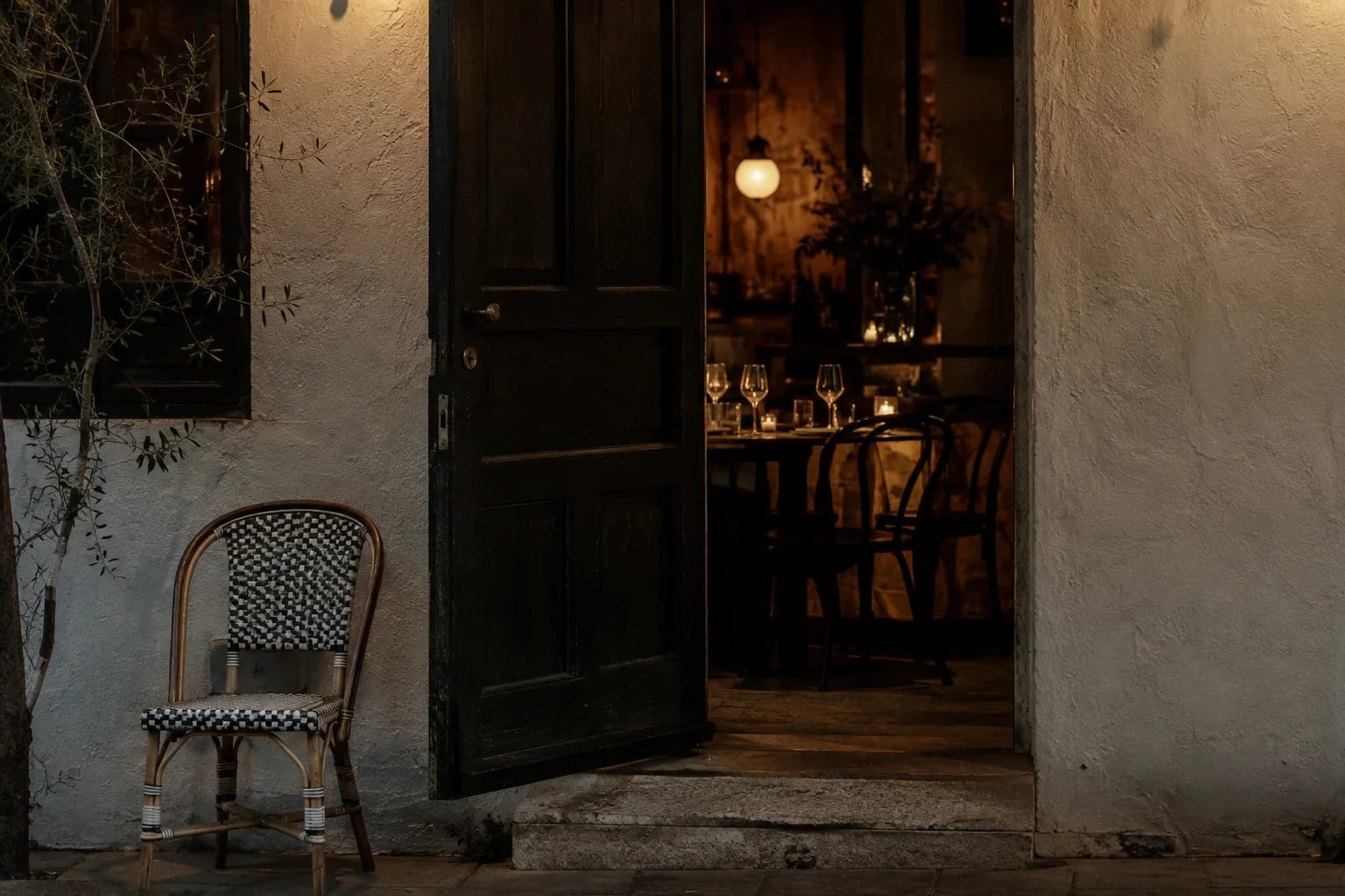 An independent neighborhood restaurant entrance at dusk, dark oak door open onto warm dining light, white plaster wall, a single empty bistro chair outside, no signage.